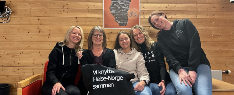 Bildet viser fem smilende personer som sitter sammen i en sofa. De holder opp en t-skjorte med teksten "Vi knytter Helse-Norge sammen".